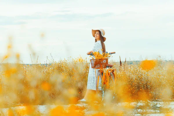 Młoda ładna kobieta ubrana w kapelusz patrząc na niebo na polu z jej żółtym rowerem. — Zdjęcie stockowe