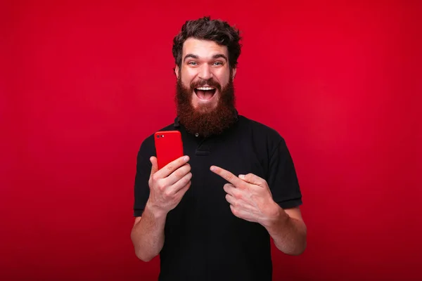 Hombre barbudo asombrado apuntando a su teléfono inteligente de pie sobre el fondo rojo —  Fotos de Stock