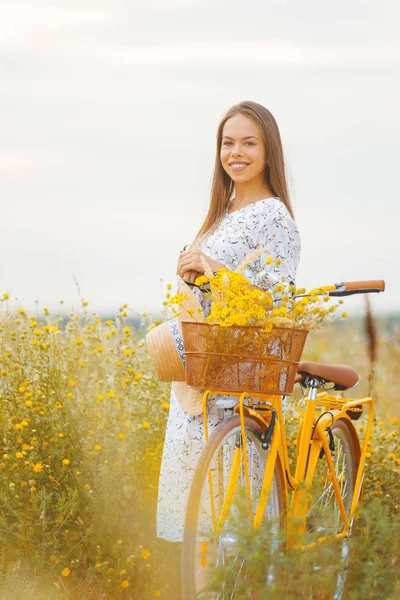 Jovencita mirando a la cámara está de pie junto a su bicicleta en un campo amarillo . — Foto de Stock