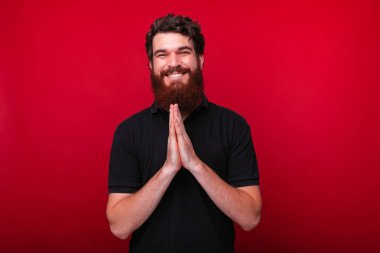 Yakışıklı sakallı adam kırmızı arka planda kameraya gülümserken dua için el ele tutuşuyor.