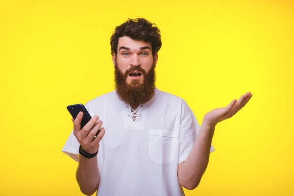 Confused fiatal szakállas férfi gazdaságban a telefon sárga háttér. — Stock Fotó