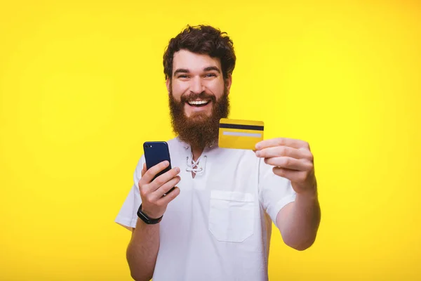 Happy internet banking customer holding his phone and yellow credit card while looking and smiling at the camera.