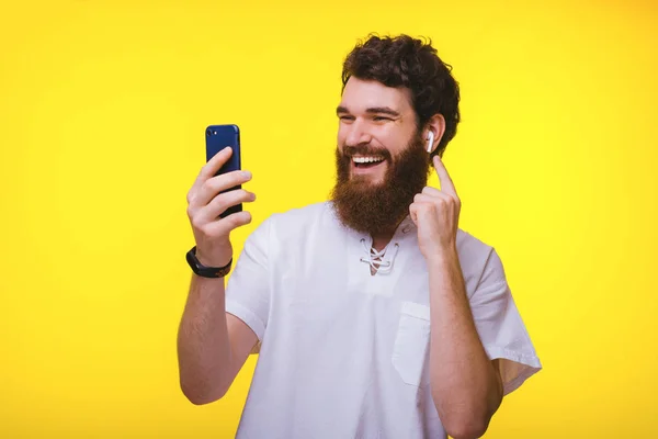 Cheerful young bearded hipster trying his new airbuds while looking at his phone on yellow background. — Stock Photo, Image