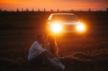 Yakışıklı çift tarlada çim üzerinde oturan, araba ışıkları üzerinde akşam