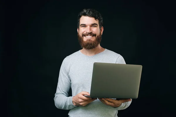 Foto de un joven sonriente sosteniendo el portátil y mirando a la cámara sobre un fondo oscuro —  Fotos de Stock