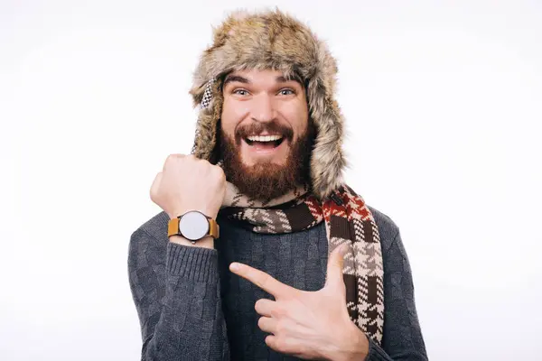 Podekscytowany brodaty mężczyzna w zimowym szaliku i czapce wskazuje na zegarek, patrząc w kamerę.. — Zdjęcie stockowe