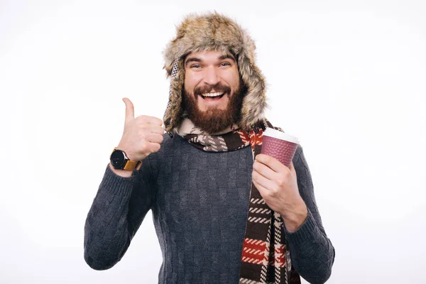 Dobra kawa Radosny brodaty hipster nosi szalik i czapkę zimową pokazując jak gest trzymając kubek na wynos. — Zdjęcie stockowe