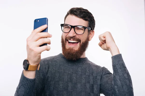 Homme barbu excité portant des lunettes est lokking à son téléphone, faire un selfie et un geste gagnant . — Photo