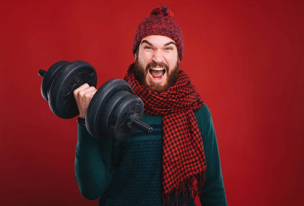 Starker bärtiger Mann in Winterkleidung hebt eine schwarze Hantel in die Höhe. Fit und gesund, Urlaubsangebot auf rotem Hintergrund. — Stockfoto