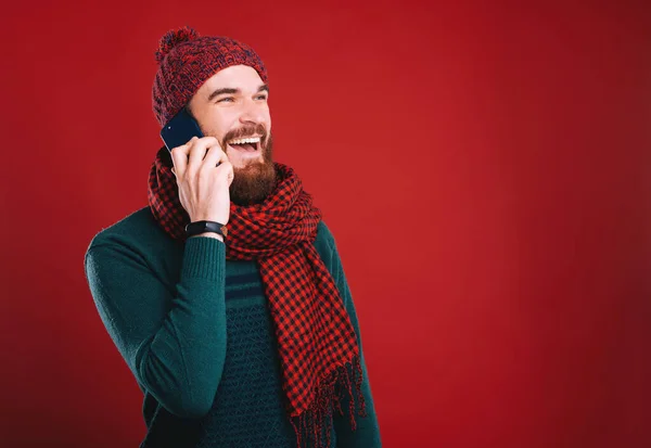 Счастливый бородатый хипстер разговаривает по телефону улыбаясь на красном фоне, одетый в теплую одежду и шарф . — стоковое фото