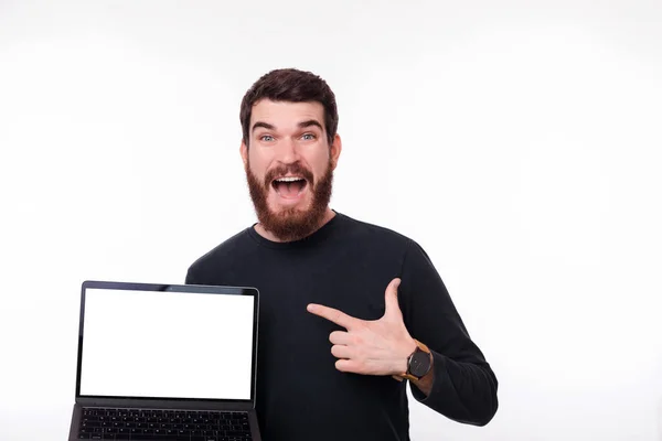 Surpris jeune homme barbu tenant son ordinateur portable et pointant vers elle sur fond blanc. Écran blanc vierge . — Photo