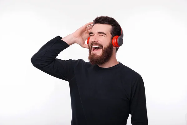 Alegre joven barbudo está escuchando música en sus auriculares, cantando sobre fondo blanco . — Foto de Stock