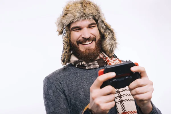 Szczęśliwy brodaty mężczyzna w czapce z futra zimowego, szalik i sweter bawiący się joystickiem, uśmiechnięty i szczęśliwy. — Zdjęcie stockowe