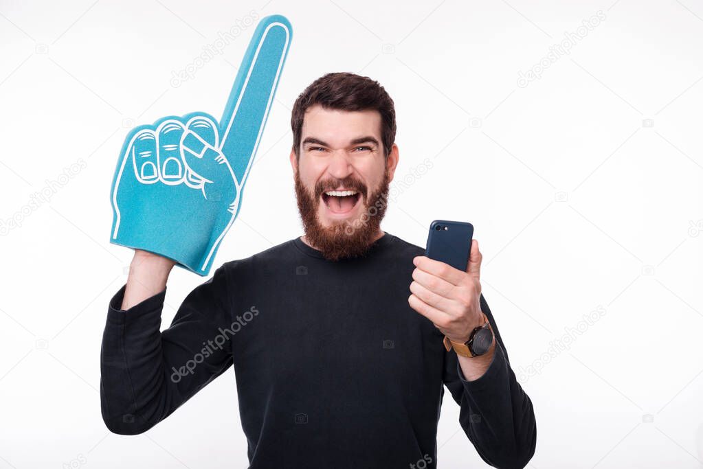 Screaming bearded man is holding his phone and fan foam finger glove