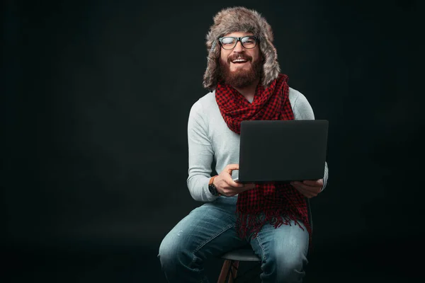 Fröhlicher bärtiger Mann sitzt auf einem Stuhl und hält seinen Laptop und lächelt in die Kamera. — Stockfoto