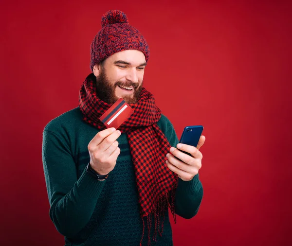 Happy bearded man using web banking, holding his card and phone.