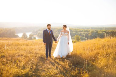 Bir gelin ve damat gün batımında bir tarlada yürüyorlar, güneş ışığında, el ele tutuşuyorlar..