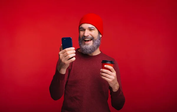 Foto de hombre barbudo sonriente en rojo mirando el teléfono y sosteniendo la taza de café — Foto de Stock