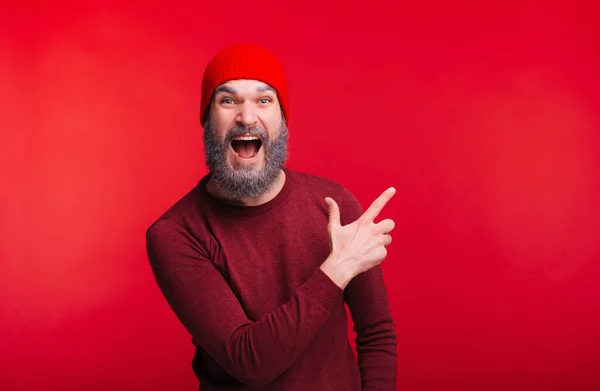 Amazed man with white beard in winter cap pointing away at copyspace over red background