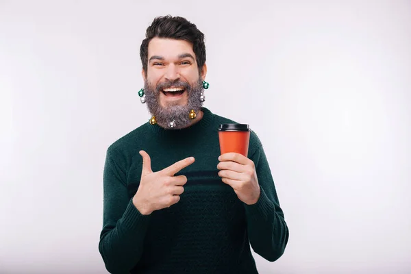Cheerful young man with decorated beard in christmas mood pointing at coffee cup to go — Zdjęcie stockowe