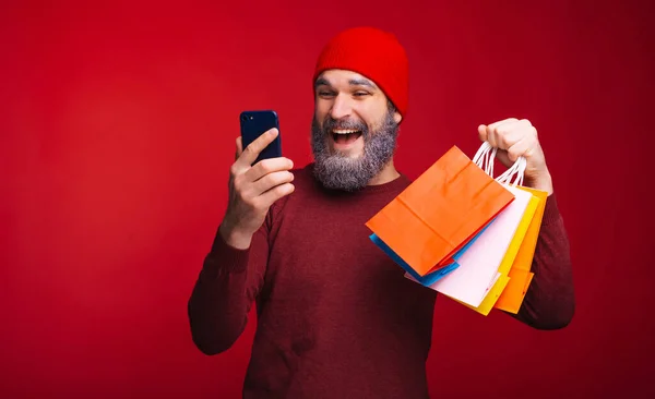 Jovem homem feliz com barba branca fazendo compras on-line e segurando alguns sacos de compras de papel — Fotografia de Stock