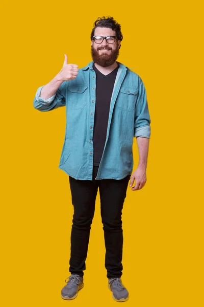 Pełna długość zdjęcie młodego brodatego mężczyzny stojącego nad żółtym tłem i pokazującego kciuki w górę — Zdjęcie stockowe