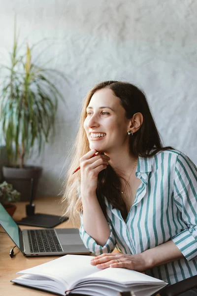 Gorgeous young woman is thinking about what to write in her planner while sitting at her work desk with plants — Stock Photo, Image