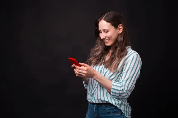Menina sorridente feliz está olhando para o telefone digitando algo no fundo preto — Fotografia de Stock