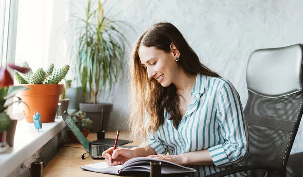 Smiling young woman is writing in her journal at home or office at work desk — Stock Photo, Image