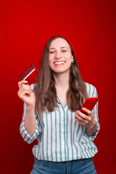 Menina sorridente vestindo camisa branca está segurando um telefone e um cartão sobre fundo vermelho — Fotografia de Stock