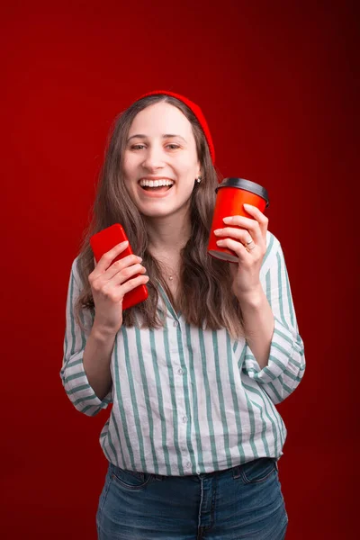 Riendo joven mujer está sosteniendo un papel para ir taza y su teléfono sobre fondo rojo — Foto de Stock