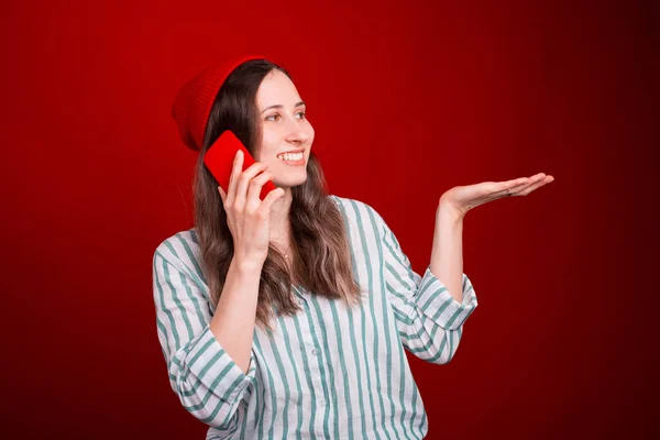 Jonge glimlachende vrouw is praten aan de telefoon, terwijl het houden van de hand omhoog presenteren iets over rode achtergrond — Stockfoto