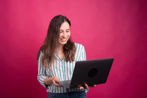 Jovem mulher está olhando animado para seu laptop enquanto está de pé sobre fundo rosa em um estúdio — Fotografia de Stock