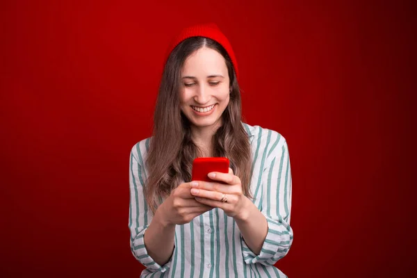 Sorrindo menina está segurando um telefone digitando sobre ele sobre fundo vermelho — Fotografia de Stock