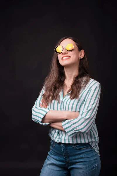 Menina sorridente confiante usando óculos de sol está de pé com os braços cruzados sobre fundo preto — Fotografia de Stock