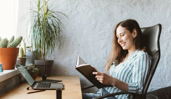 Lächelnde junge Frau schaut in ein Buch oder einen Planer, während sie auf ihrem Schreibtisch mit Pflanzen sitzt — Stockfoto