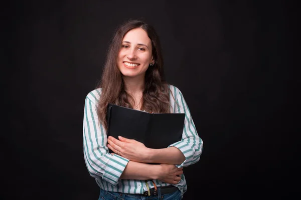 Linda joven mujer abraza un libro abierto mientras sonríe a la cámara sobre fondo negro — Foto de Stock