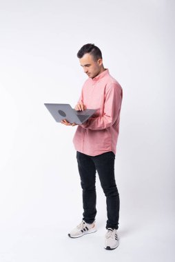 Beyaz arka planda tuttuğu dizüstü bilgisayarı kullanan genç bir adamın tam vücut fotoğrafı.