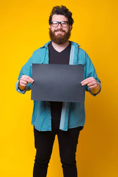 对着摄像机微笑的男人拿着一张站在黄色背景上的黑纸 — 图库照片