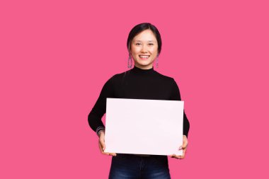 Kameradaki kadına gülümseyen kadın, elinde mesajın için beyaz bir tahta tutuyor, pembe arka planda duruyor.