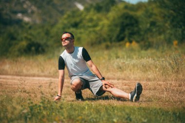 Güneş gözlüğü takan genç adam bir tarlada ısınma egzersizleri yapıyor..