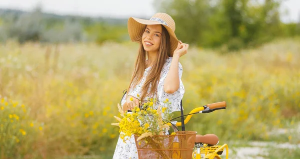 Retrato de una mujer joven con un sombrero, está de pie cerca de su bicicleta en un campo en verano — Foto de Stock