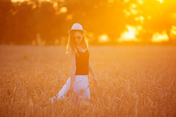 Junge Frau mit Kleid und Hut geht an einem sonnigen Tag durch ein Weizenfeld — Stockfoto