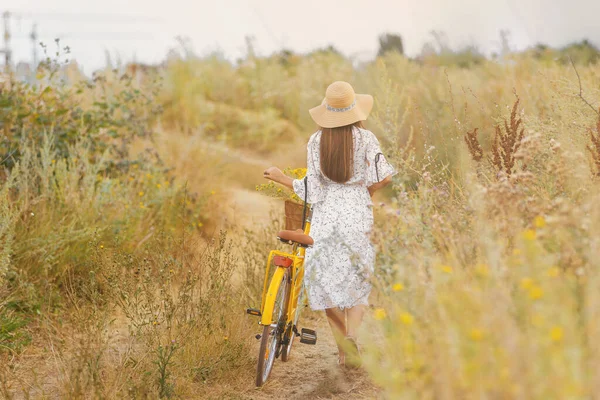Foto de una joven vestida de blanco y un sombrero caminando con su bicicleta amarilla en un campo — Foto de Stock