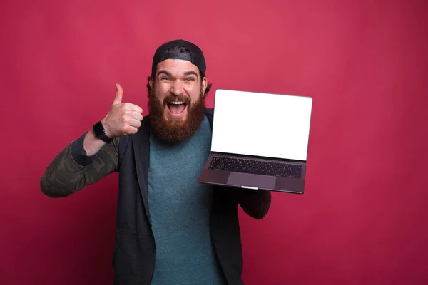 Excited bearded man is holdinghis laptop with blank screen and showing thumb up or like gesture over pink background.