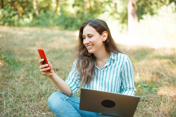 Foto de mulher bonita feliz usando seu telefone no parque enquanto sentado na grama — Fotografia de Stock