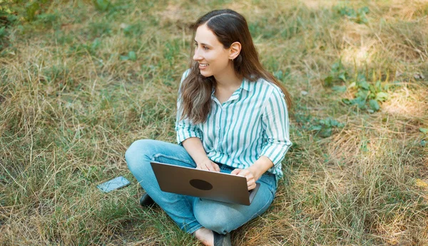 Foto av glad ung kvinna sitter på gras i parken och använder laptop — Stockfoto