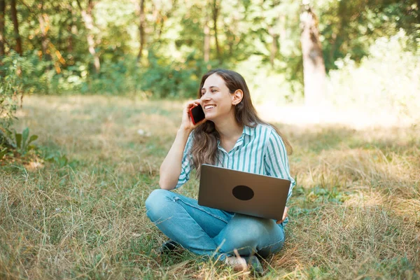 Foto av leende och charmig kvinna talar i telefon medan du sitter på gräs i parken — Stockfoto