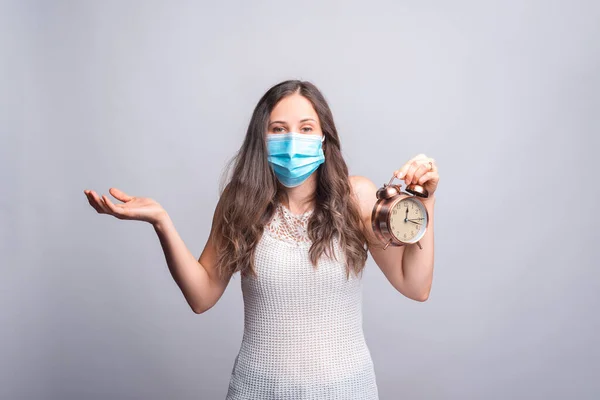Retrato de mulher jovem usando máscara medicinal e segurando despertador sentindo confuso — Fotografia de Stock