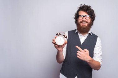 Takım elbiseli genç adam beyaz arka planda çalar saati işaret ediyor..